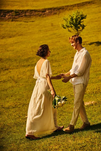 redhead groom and asian bride with flowers wearing elegant boho attire and holding hands in field