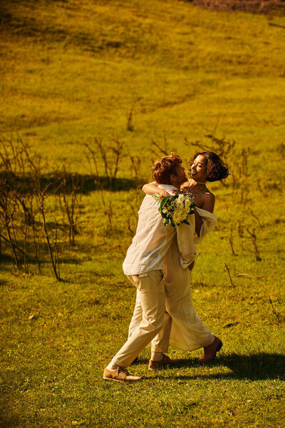 redhead groom embracing cheerful asian bride with flowers in green field, rural celebration