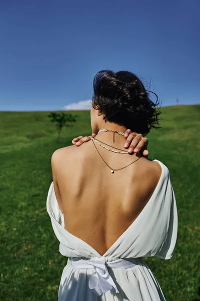 young brunette woman in necklaces and wedding dress with naked back under blue sky in scenic field