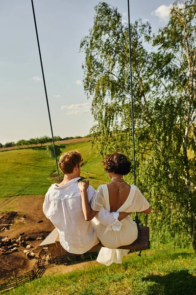 stock image rustic wedding, back view of redhead groom with young bride swinging in picturesque countryside