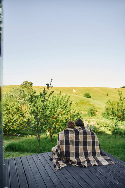 back view of young couple sitting on wooden porch under plaid blanket and enjoying scenic landscape