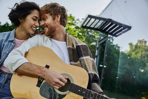 happy redhead man sitting face to face with asian girlfriend and playing guitar near glass house