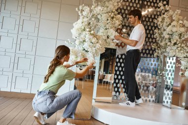Gülümseyen adam ve genç kadın modern etkinlik salonunda beyaz çiçek dekoru düzenliyorlar, yaratıcı takım çalışması.