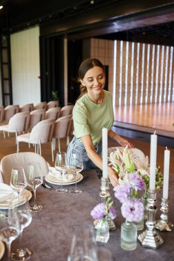 Genç dekoratör, kutlama salonunda şenlik ortamı, çiçekler ve mumlarla masanın yanında gülümsüyor.