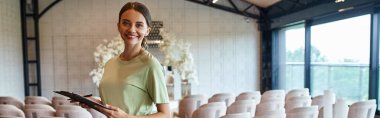 joyful event coordinator with clipboard smiling at camera in decorated banquet event hall, banner clipart