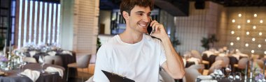 Panosu olan mutlu etkinlik yöneticisi şölen salonundaki şenlik masalarının yanında akıllı telefondan konuşuyor.