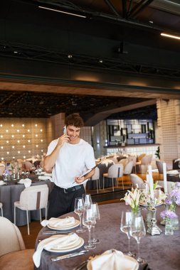 smiling event coordinator with clipboard talking on smartphone near festive tables in banquet hall clipart