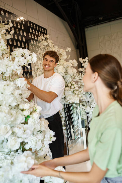 smiley decorator arranging flower decor in event hall and looking at colleague on blurred foreground