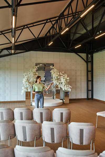 молодой организатор мероприятия с буфером обмена, осматривающий зал мероприятий с цветочным декором, праздничной обстановкой