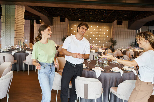 cheerful team lead with clipboard pointing with hand near happy team in modern banquet hall