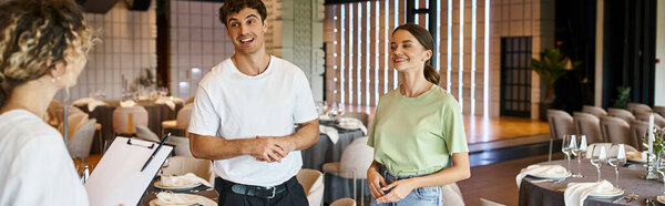 позитивные менеджеры мероприятия, улыбающиеся рядом с командными и праздничными столами в украшенном банкетном зале, баннере