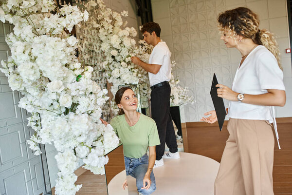 happy woman looking at team lead with clipboard while working with floral decor in event hall
