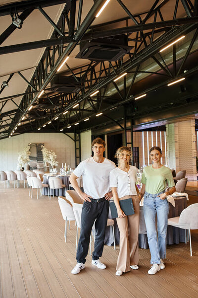 довольная команда организаторов мероприятия, смотрящая на камеру возле праздничных столов в украшенном банкетном зале