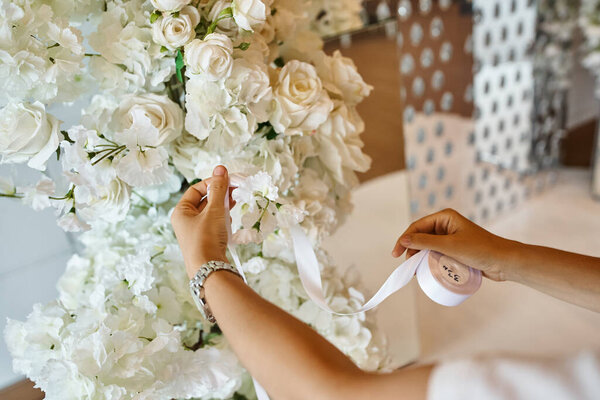 cropped view of decorator holding white ribbon near blooming flowers in event hall, banquet setup