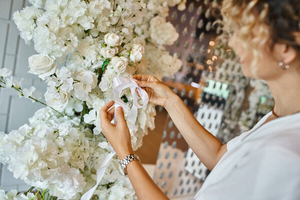 creative florist tying white ribbon on blooming floral composition in event hall, banquet setting