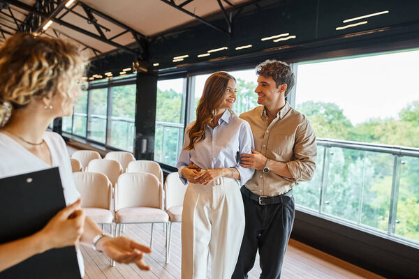 joyful couple smiling at each other near banquet coordinator with clipboard in modern event hall