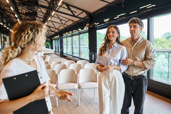 smiling couple in love looking at event organizer with clipboard talking in modern wedding venue