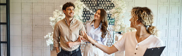 banquet coordinator with clipboard showing event hall with floral decor to happy couple, banner