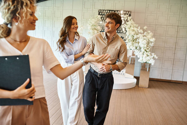 joyful couple in love smiling near banquet organizer showing event hall with wedding decoration