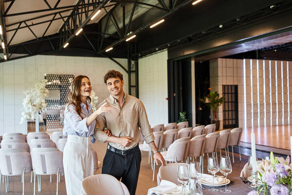 amazed woman pointing with hand near festive table and boyfriend in event hall with wedding decor