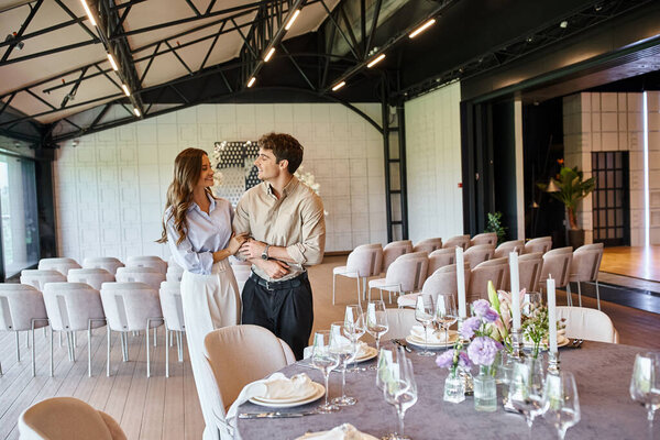 happy couple looking at each other near festive table decorated with flowers in modern wedding venue