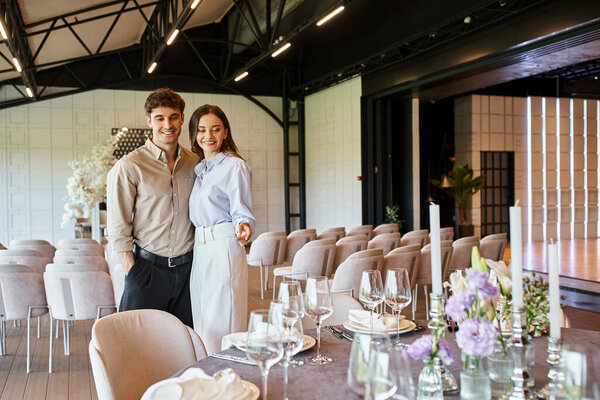 pleased woman pointing at festive table with wedding decor near happy boyfriend in event hall