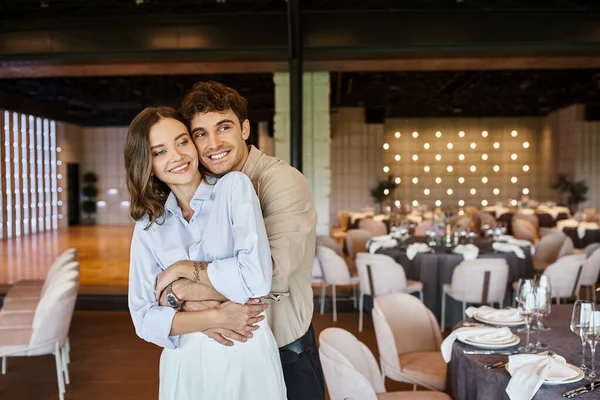 stock image overjoyed man embracing girlfriend in banquet hall with decorated festive tables, wedding setup