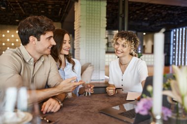 smiling man talking to event manager near happy girlfriend in banquet hall, wedding preparation clipart