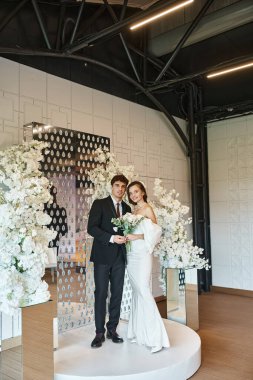 Tam boy romantik yeni evli çift etkinlik salonunda beyaz çiçeklerle süslenmiş şekilde poz veriyorlar.