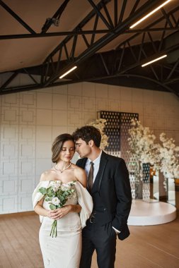 Beyaz gelinlik içinde büyüleyici bir gelin, siyah takım elbiseli damadın yanında çiçeklerle dolu bir salonda.