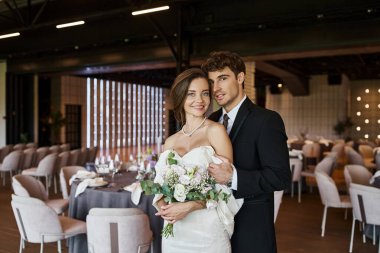 Modern dekore edilmiş ziyafet salonundaki kameraya gülümseyen gelin buketiyle mutlu ve zarif yeni evliler.