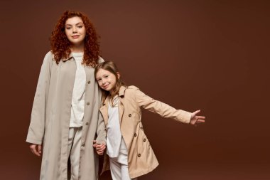 Modern ebeveynlik, kıvırcık anne kızıyla el ele tutuşuyor ve kahverengi arka planda paltoların içinde duruyor.