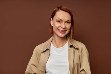 happy redhead woman in beige trench coat looking at camera on brown backdrop, autumn fashion clipart