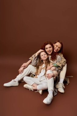 three generations, cheerful redhead family with freckles looking at camera on brown background clipart