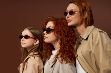 three female generations with red hair posing in sunglasses and coats on brown backdrop, family bond clipart