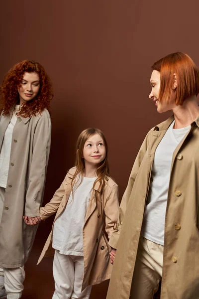 three generations, happy redhead family in coats holding hands on brown backdrop, women and girl