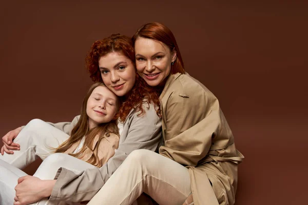 three generations of women, positive redhead family looking at camera on brown background, autumn