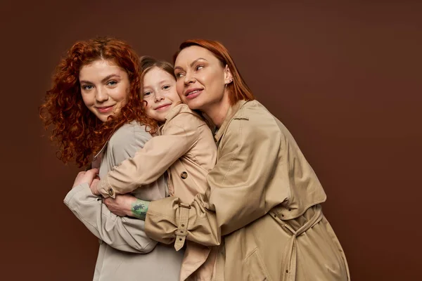 joyful redhead family of three female generations hugging on brown background, autumn fashion