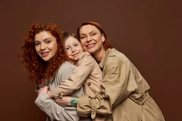 happy redhead family of three female generations hugging on brown background, autumn fashion