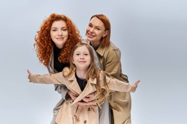 Gri arka planda bej renkli sonbahar ceketleriyle poz veren kızıl saçlı kadınlar ve çocuklar.