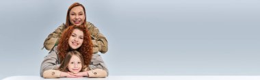 Üç nesil, gri arka planda poz veren bej renkli trençkotlu kızıl saçlı anne ve çocuk, afiş.