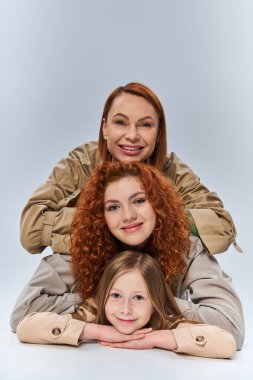 happy three generations, redhead mother and child in beige trench coats posing on grey background clipart