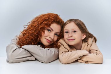 Mutlu iki nesil, gri arka planda gülümseyen bej trençkotlu kızıl saçlı anne ve çocuk.