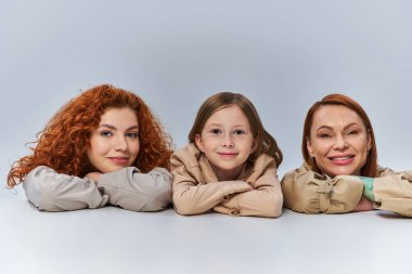 joyful female generations, redhead women and child in beige coats smiling on grey background, family clipart