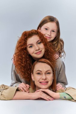 three female generations, happy redhead family in beige coats smiling on grey backdrop, family bond clipart