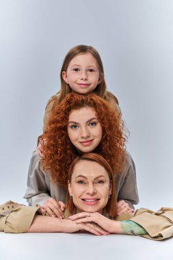 three generations, happy redhead family in beige coats smiling on grey backdrop, female bond clipart