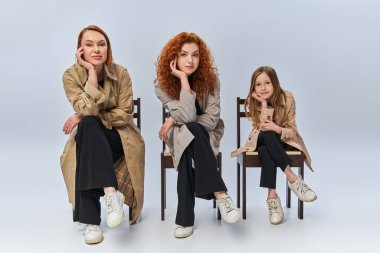 Üç kadın nesil sırayla sandalyede oturuyor, mutlu kızıl saçlı kadınlar ve gri arka planda çocuk