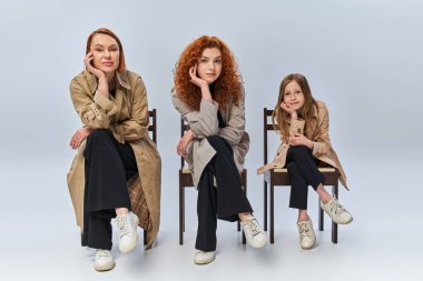 three female generations sitting in row on chairs, happy redhead women and child on grey backdrop clipart