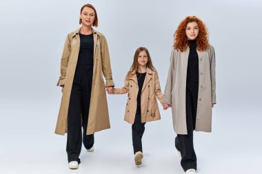 three female generations, redhead women and girl in coats holding hands and walking on grey backdrop clipart