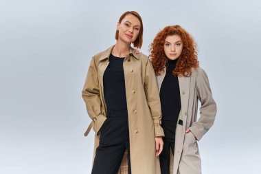 two generations of redhead women posing in coats and looking at camera on grey background, family clipart